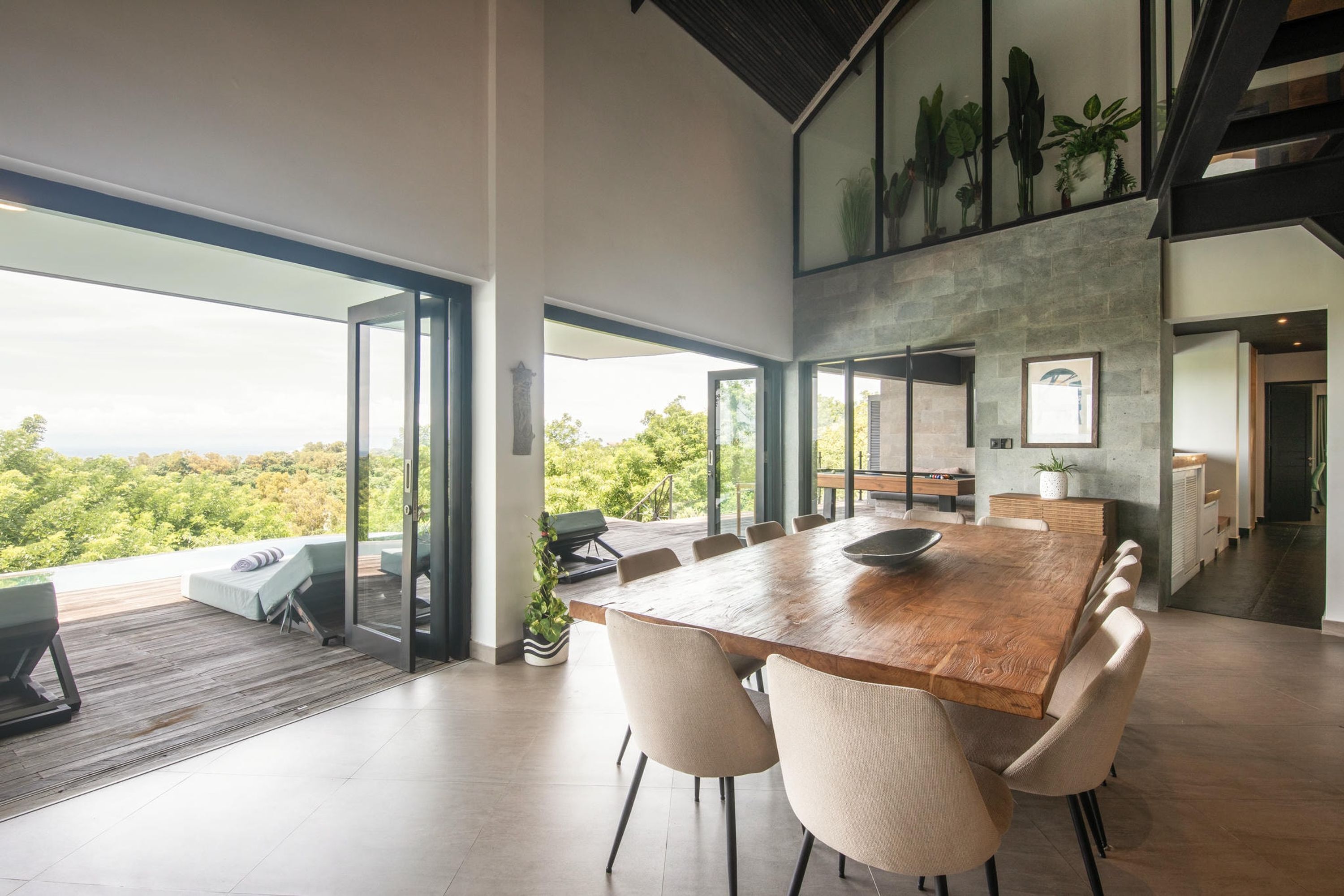 Un comedor amplio con una gran mesa de madera y múltiples sillas, ventanas panorámicas que ofrecen vistas al exterior, y un diseño arquitectónico moderno con una pared y techo de piedra.