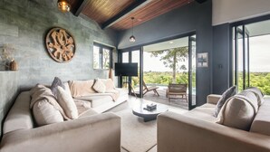 Ein modernes Wohnzimmer mit einer großen Couch, einem Couchtisch und einem Fernseher. Es gibt eine Holzbalkendecke und große Fenster mit Blick auf Grünflächen.