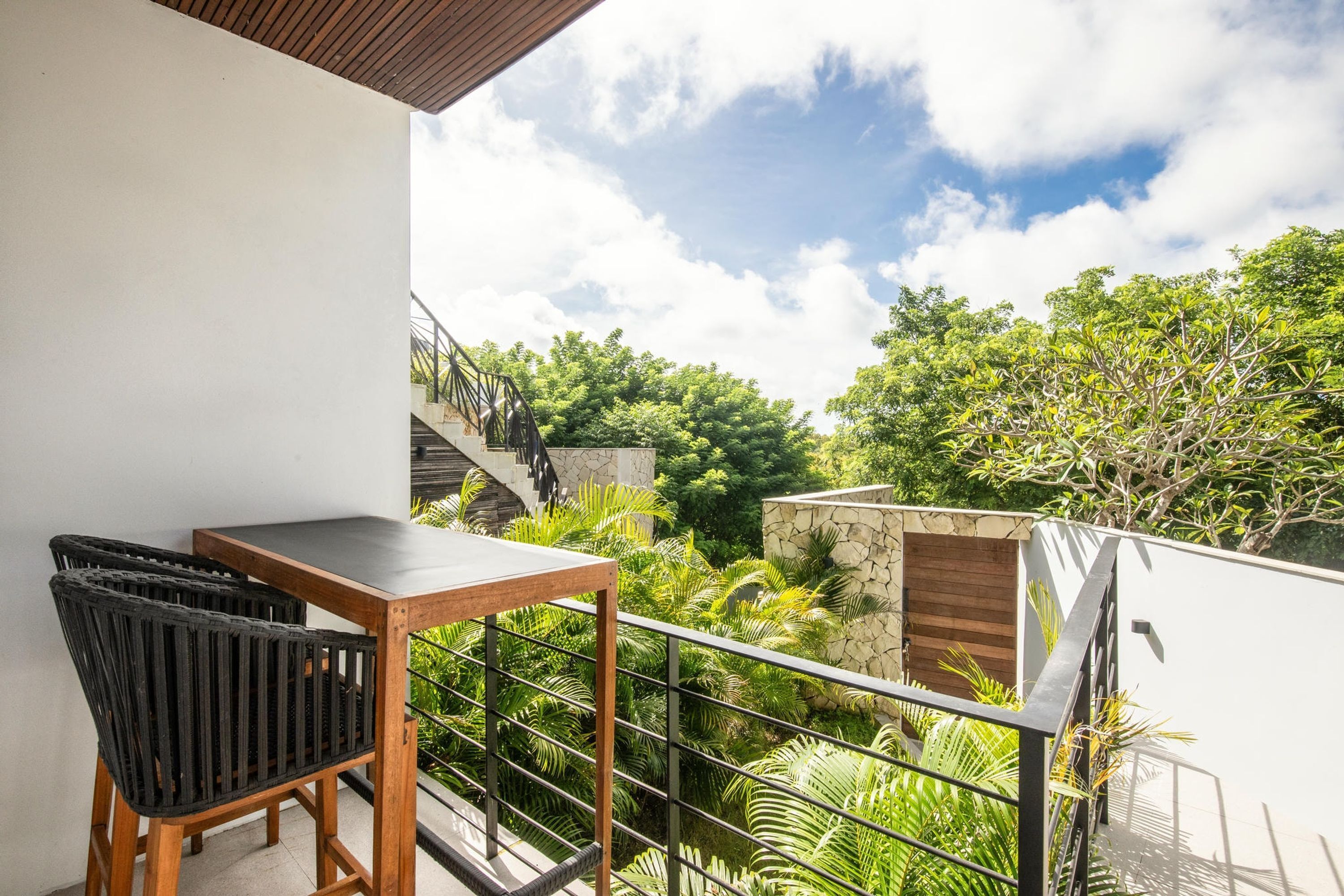A balcony with a wooden table and chairs, a stone wall, and lush greenery in the background.