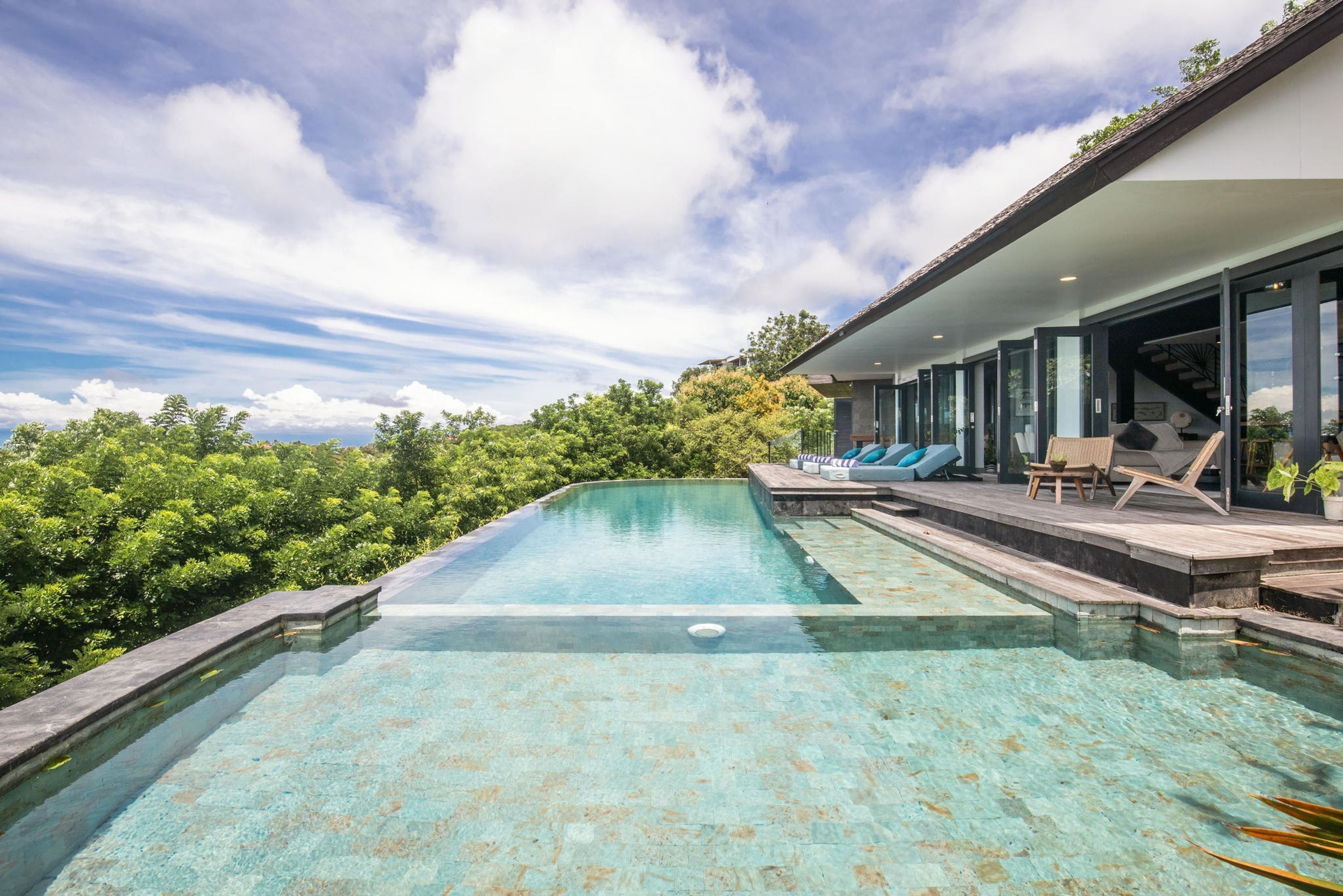 Ein moderner Infinity-Pool mit klarem Wasser, umgeben von üppiger Vegetation und einem großzügigen Haus mit überdachter Terrasse.