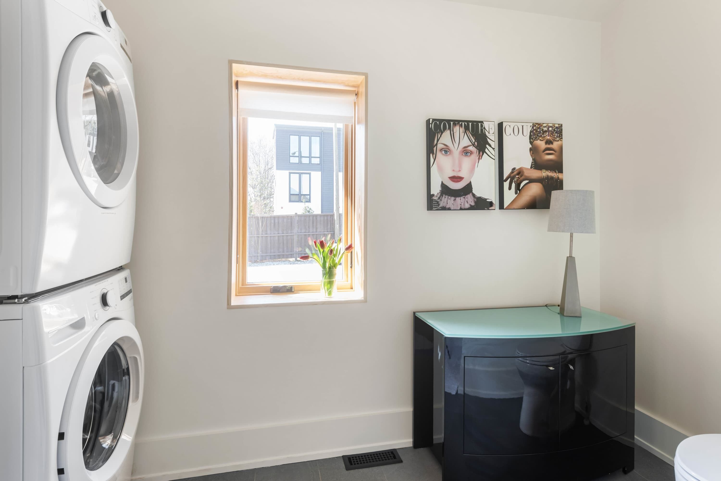 A laundry area with a washing machine, a window with a view of a building, and a cabinet with a lamp.