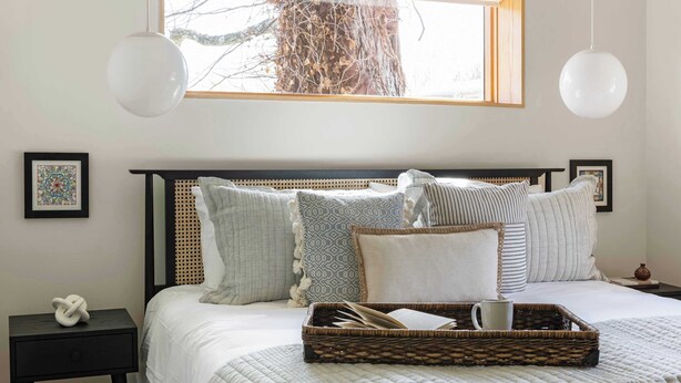 A neatly made bed with a quilted blanket, a wicker tray with a book and a mug, and a framed picture on the nightstand.
