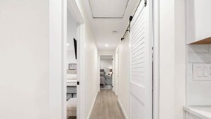 A narrow hallway with a wooden floor, white walls, and a door leading to a bedroom with a bed and a framed picture on the wall. - Freshly Updated Hideaway in CDA’s Best Spot (Coeur d'Alene)