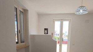 Un salon avec un canapé beige, une fenêtre avec des stores, une porte donnant sur l’extérieur et un luminaire suspendu.