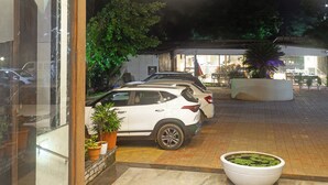 Entrada de un hotel con puerta de cristal, un coche blanco estacionado afuera y una jardinera con plantas verdes.