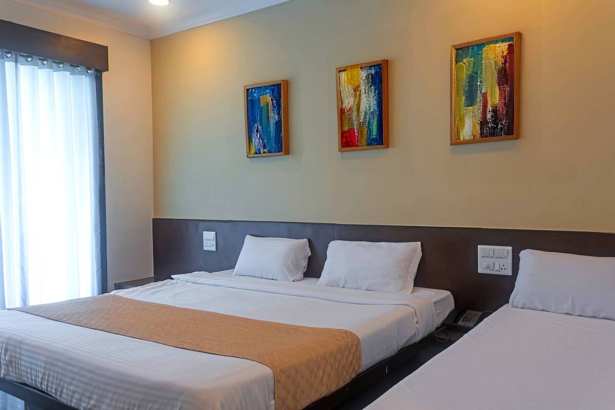A hotel room with two beds, a large window with curtains, and three framed abstract paintings on the wall.