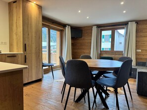 A modern kitchen with wooden cabinets, a dining table with chairs, and large windows offering a view of the outdoors.