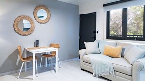 Uma sala de estar moderna com uma mesa branca e duas cadeiras de vime, um sofá bege com um cobertor, e dois espelhos fixados na parede.