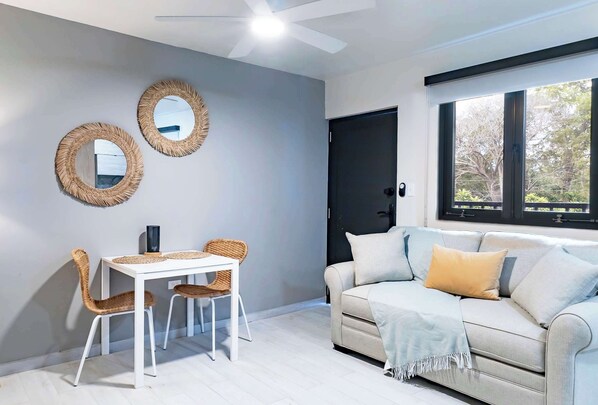 Una sala de estar moderna con una mesa blanca y dos sillas de mimbre, un sofá color beige con una manta, y dos espejos colgados en la pared.