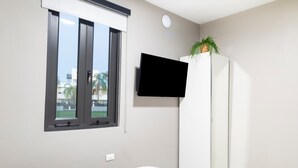 Un dormitorio con una cama, un televisor montado en la pared, una ventana con vistas a palmeras y una planta sobre un mueble.