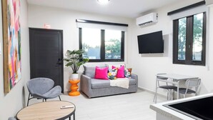 Una sala de estar moderna con un sofá gris, cojines coloridos, una planta en maceta, una mesita redonda y un televisor montado en la pared.