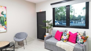 Un salón moderno con un sofá gris decorado con almohadones de colores, un sillón gris, una mesita redonda y un gran ventanal con vistas a palmeras y un estacionamiento.