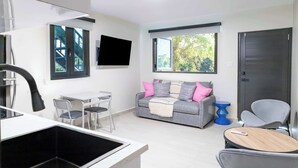 Una sala de estar moderna con un sofá gris, un televisor montado en la pared, un comedor con mesa y sillas, y un ventanal con vistas a la vegetación.