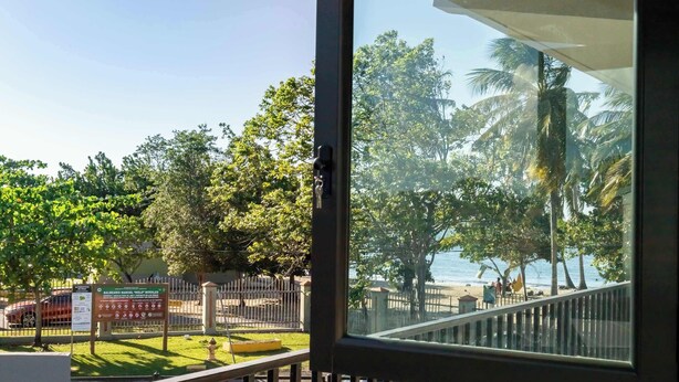 Vista desde una ventana que da a una playa con palmeras, una valla y un cartel.