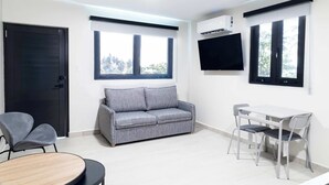 En moderne stue med en grå sofa, et lille spisebord og to stole. Der er et tv ophængt på væggen og et vindue med persienner.