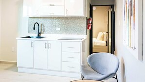 Et moderne køkken med hvide skabe, et backsplash i fiskebensmønster og en sort vandhane. Der er et rundt træbord med to stole, og en gang der fører til et soveværelse.