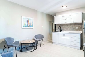 A modern kitchen with white cabinets, a black faucet, and a herringbone-patterned backsplash. There is a round table with two chairs and a painting of sailboats on the wall.