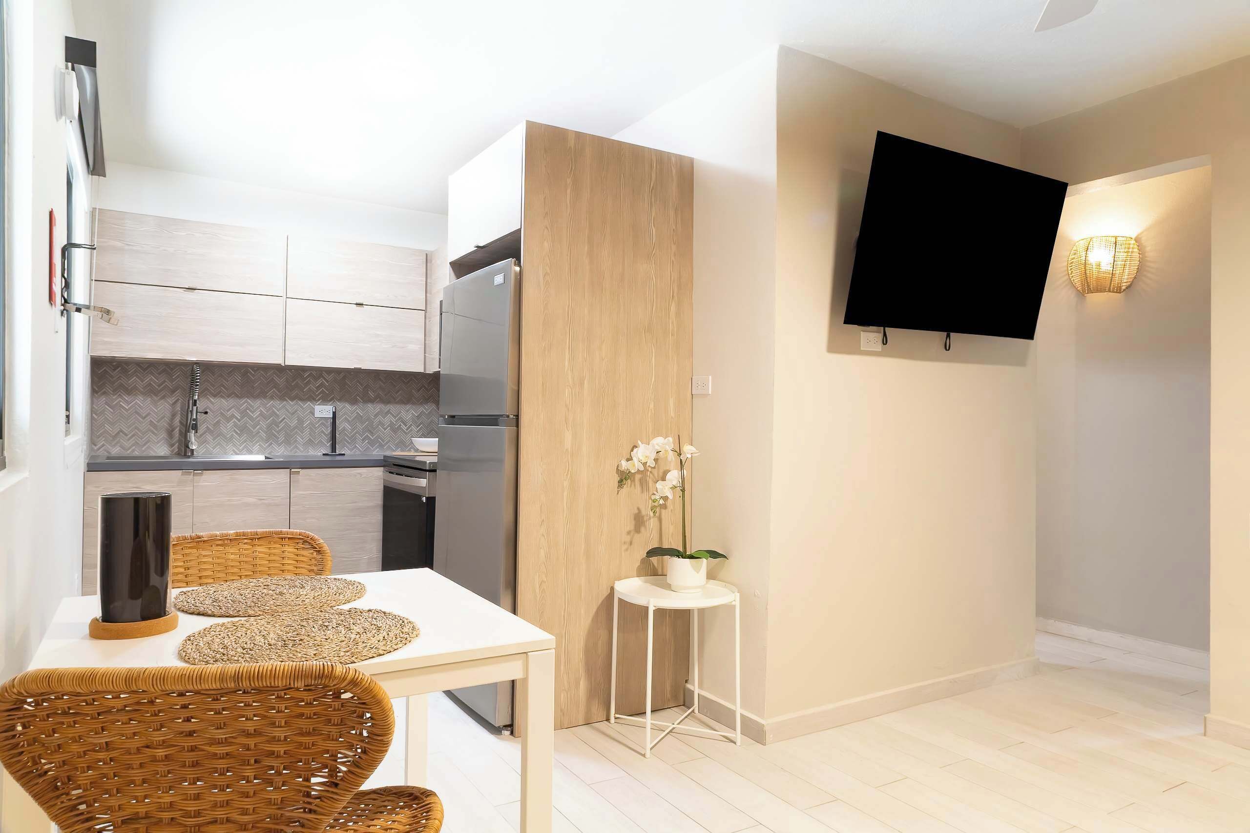 Une cuisine moderne avec des meubles de couleur bois clair, un four encastré et un réfrigérateur. On y trouve un coin repas avec une table blanche et des chaises en osier. Une petite table d’appoint blanche, sur laquelle est posée une orchidée en pot, se trouve à côté de la table à manger. Un téléviseur à écran plat est fixé au mur, au-dessus du coin repas.