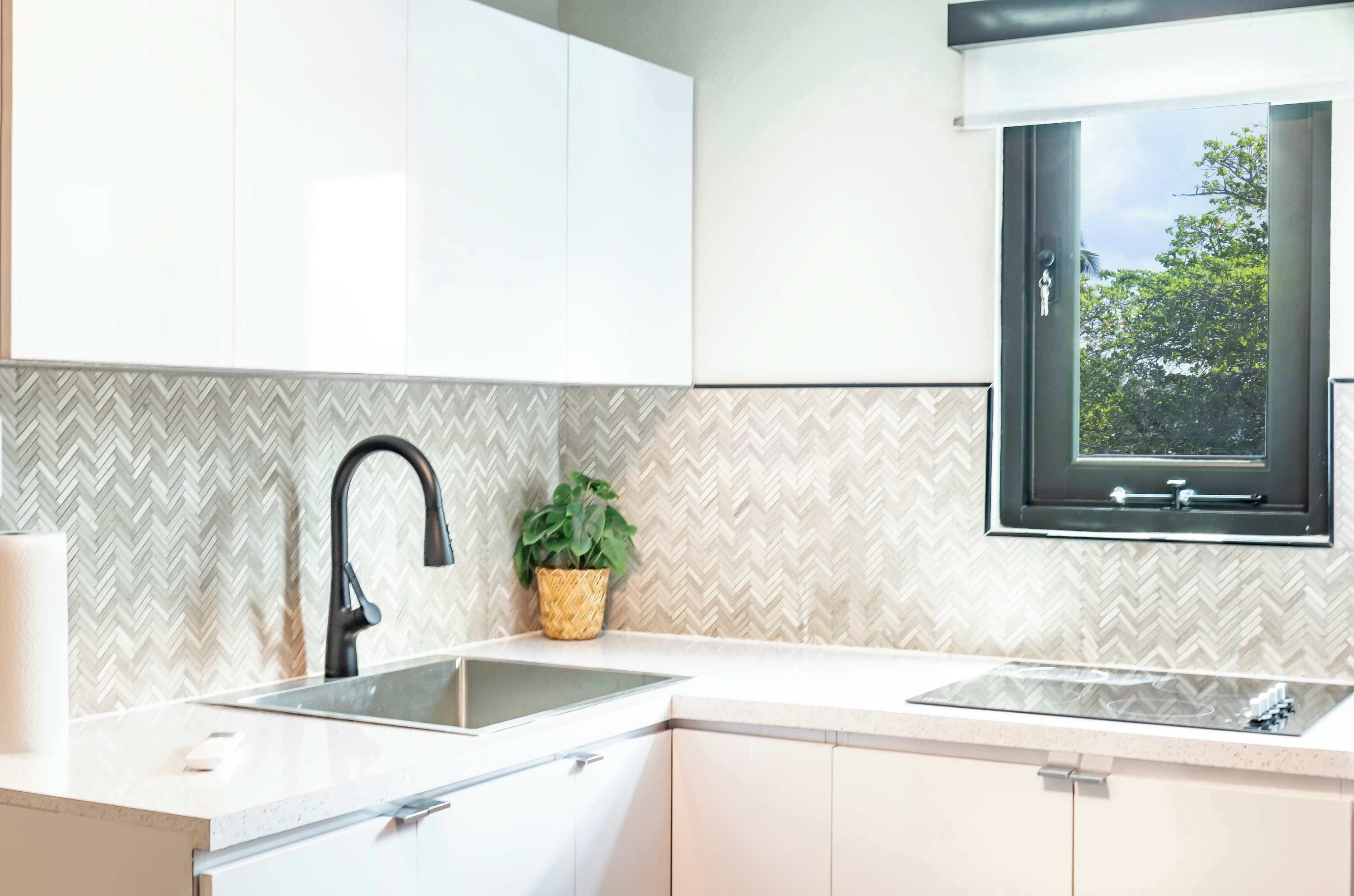 A modern kitchen with white cabinets, a herringbone-patterned backsplash, a black faucet, a window with a view of greenery, and a small potted plant on the counter.