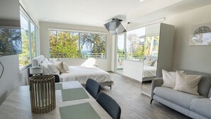 A modern hotel room with a large bed, a sofa, and a dining table. There are large windows providing a view of the outdoors. - AQUA Terrazas 6+7 · 3BR Fun & Dining by the Beach (Dorado)