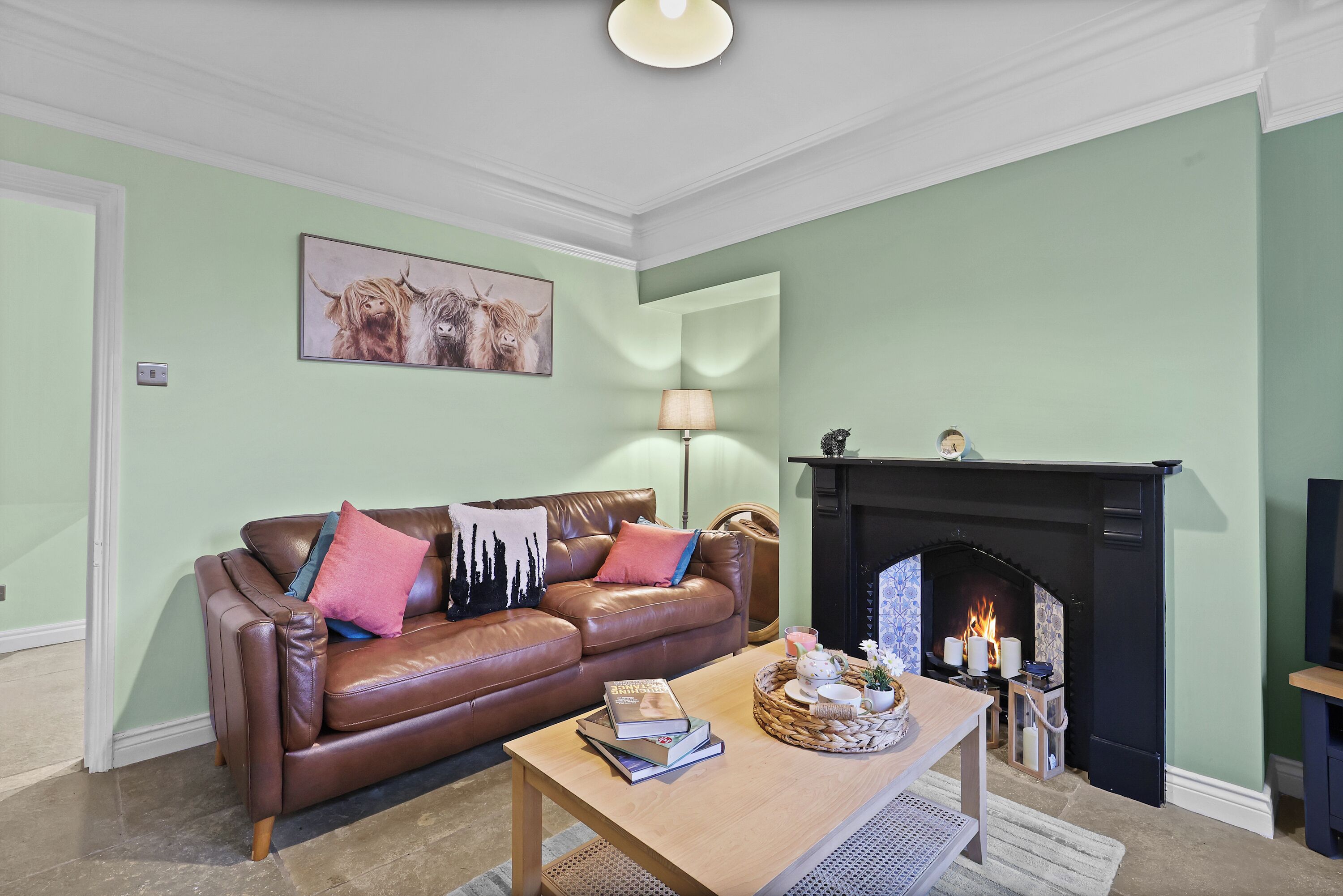 A living room with a brown leather sofa, a wooden coffee table with books and decorative items, a fireplace, and a painting on the wall.
