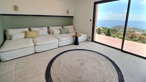 Un salon moderne avec un grand canapé, une table basse et une vue sur la mer, visible à travers une porte-fenêtre coulissante.