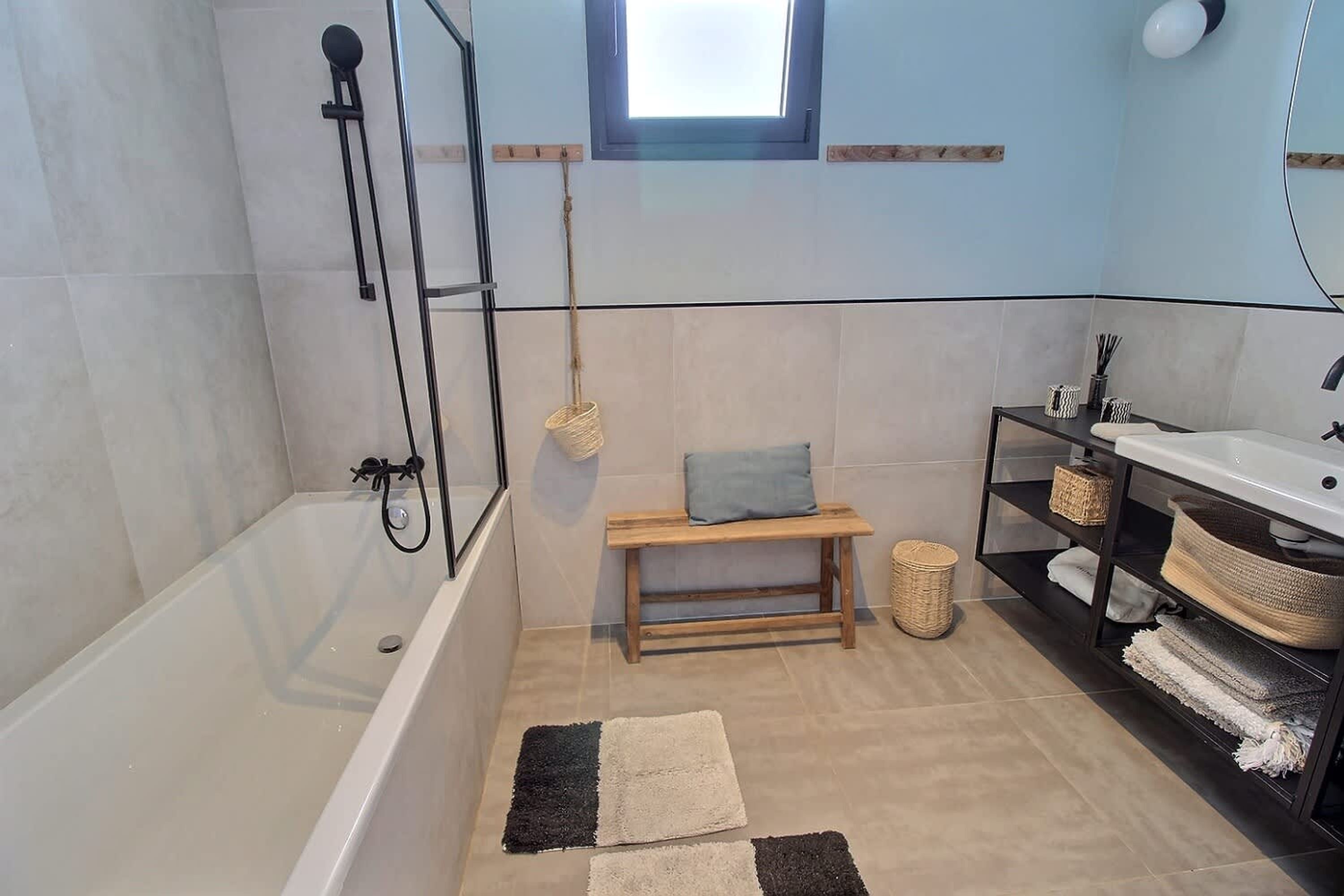 Un baño moderno con bañera, una banca de madera, un estante con cestas y una ventana pequeña.