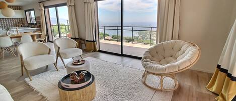 A spacious living room with a large window offering a view of the sea, a dining area with a table and chairs, and a cozy seating area with a round table and cushions.