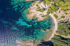 An aerial view of a coastal area with clear turquoise waters, rocky terrain, and a small building with a roof.