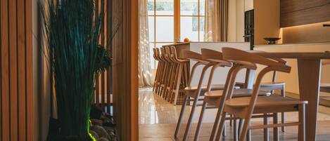A modern dining area with wooden chairs and a central table, a large potted plant, and a view through a glass door to a kitchen area.
