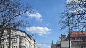 Egy tágas tér, központi gyalogosátkelővel, melyet történelmi épületek vesznek körül, a távolban pedig egy piros busz látható.
