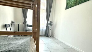Egy szoba, amelyben van egy ágy, egy íróasztal, egy szék, egy ventilátor és egy kép a falon.