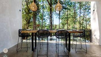 An outdoor dining area with a long wooden table and four black chairs, surrounded by trees and a glass railing.