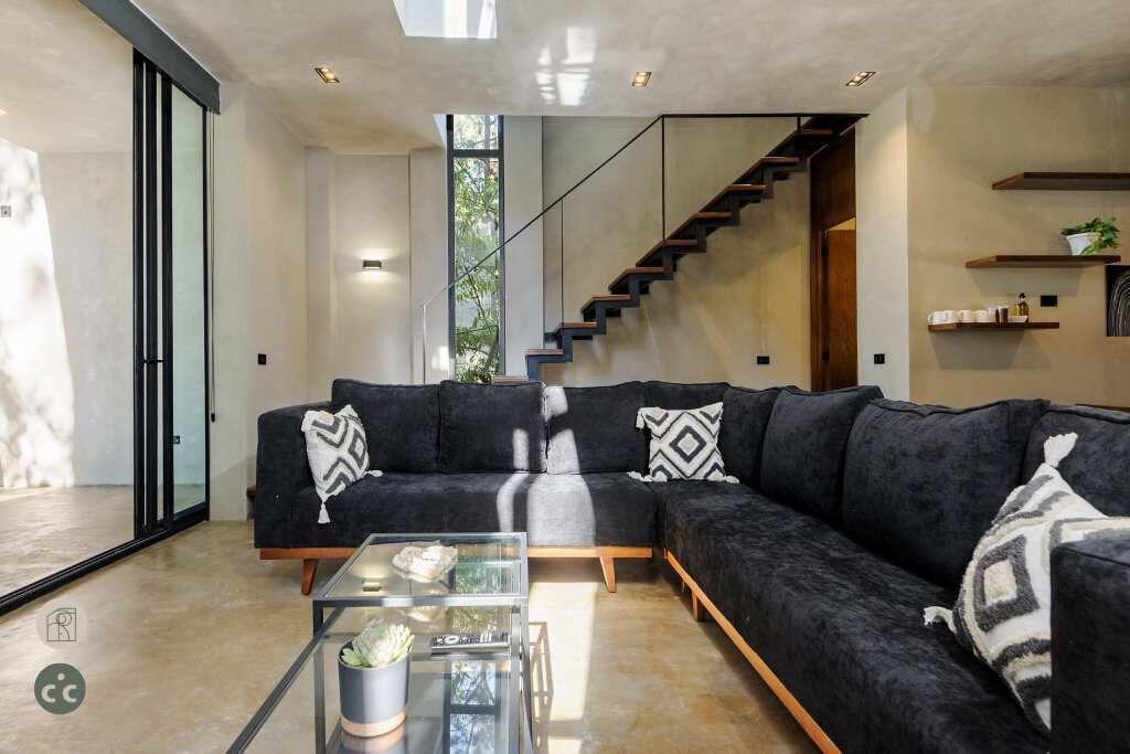 A modern living room with a black sectional sofa, a glass coffee table, and a staircase leading to an upper floor.