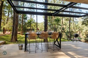 Un área de comedor al aire libre con cerramiento de vidrio, rodeada de árboles.