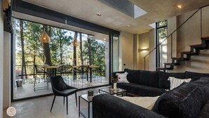 Una sala moderna con un gran ventanal que da a un espacio exterior, un sofá negro, una mesa de centro y una escalera.
