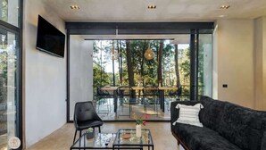 Una sala de estar moderna con un sofá negro, una mesa de centro de vidrio y una silla negra. Hay un televisor montado en la pared y un ventanal con vistas a árboles.