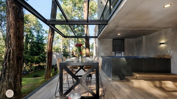 Un espacio al aire libre para comer con mesa y sillas modernas, rodeado de árboles y una estructura de techo de cristal.