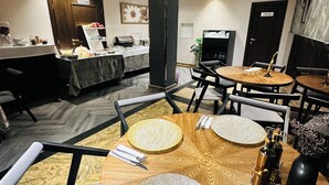 A dining area with round wooden tables, each set with a plate, cutlery, and a napkin. There are chairs around the tables, and a buffet counter is visible in the background.
