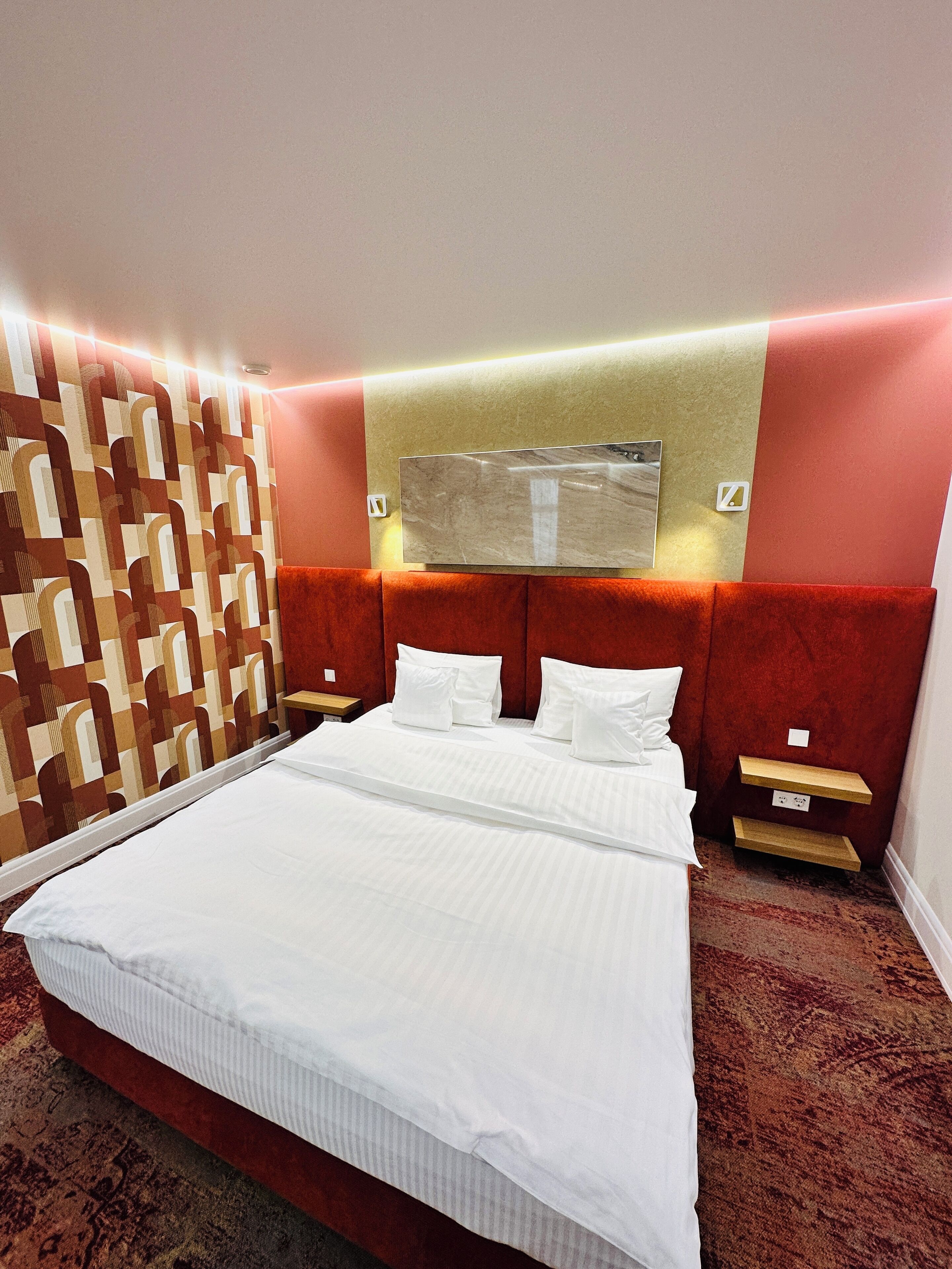 A hotel room with a large bed, two bedside tables, a patterned wall, and a headboard with integrated lighting.