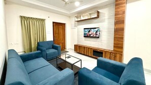 A living room with blue sofas, a coffee table, a TV mounted on a wooden wall, and a ceiling fan. - 3 Bee's Service Apartments (Hyderabad)