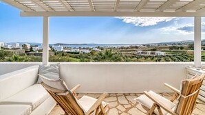 A covered outdoor seating area with a white sofa and wooden chairs, overlooking a coastal landscape with buildings and greenery. - Quiet area with Beach access (Chrisi Akti)