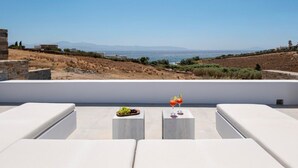 Una terraza con sofás blancos y una mesa con frutero y bebidas, con vistas a un paisaje costero.