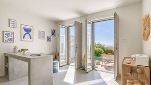 A modern kitchen with a central island, a dining area with a table and chairs, and a view of the outdoors through large sliding doors. - Spick & span in Paros island (Aspro Chorio)