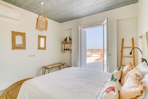 Un dormitorio con una cama grande, un banco de madera, una silla de mimbre y vistas al exterior a través de una puerta abierta.