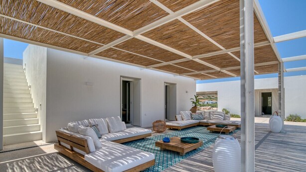 A covered outdoor seating area with a sofa and two side tables, a large white vase, and a view of the surrounding landscape.