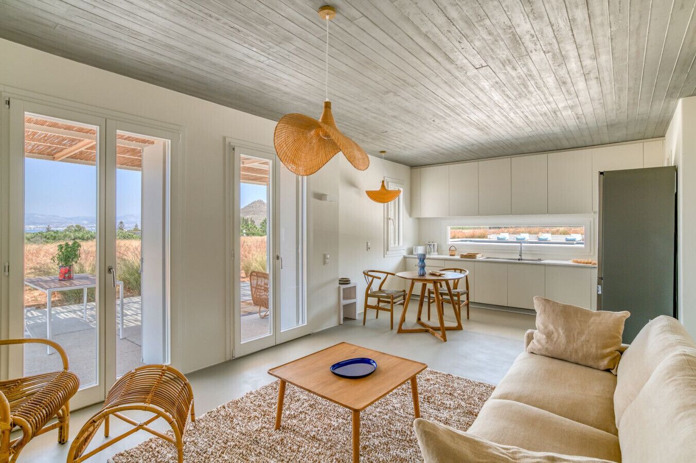 Ein modernes Wohnzimmer mit einer Couch, einem Couchtisch und Korbstühlen. Es gibt eine Küchenzeile mit einem Esstisch und Stühlen. Das Zimmer verfügt über große Schiebeglasfenster, die auf eine Außenpatio führen.