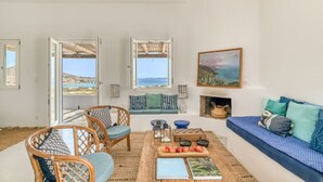 A bright living room with a view of the sea, wicker furniture, a coffee table with books, and a blue sofa with cushions. - Villa Tchi Tcha - Naoussa Bay (Kamares Naousas)
