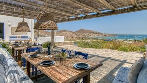 An outdoor dining area with wooden tables and chairs, a thatched roof, and a view of the sea and mountains. - Villa Tchi Tcha - Naoussa Bay (Kamares Naousas)