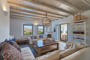A spacious living room with a wooden ceiling, a fireplace, a large window overlooking a landscape, and a bookshelf.